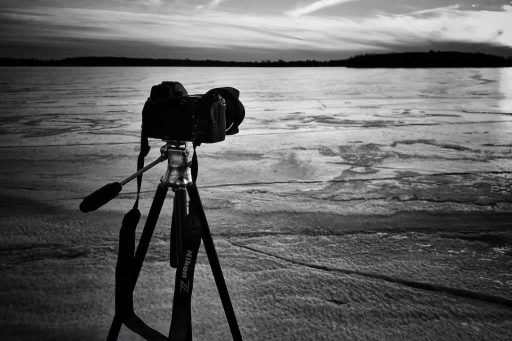 Camera on tripod capturing a serene frozen lake at dusk with dramatic sky in monochrome photography