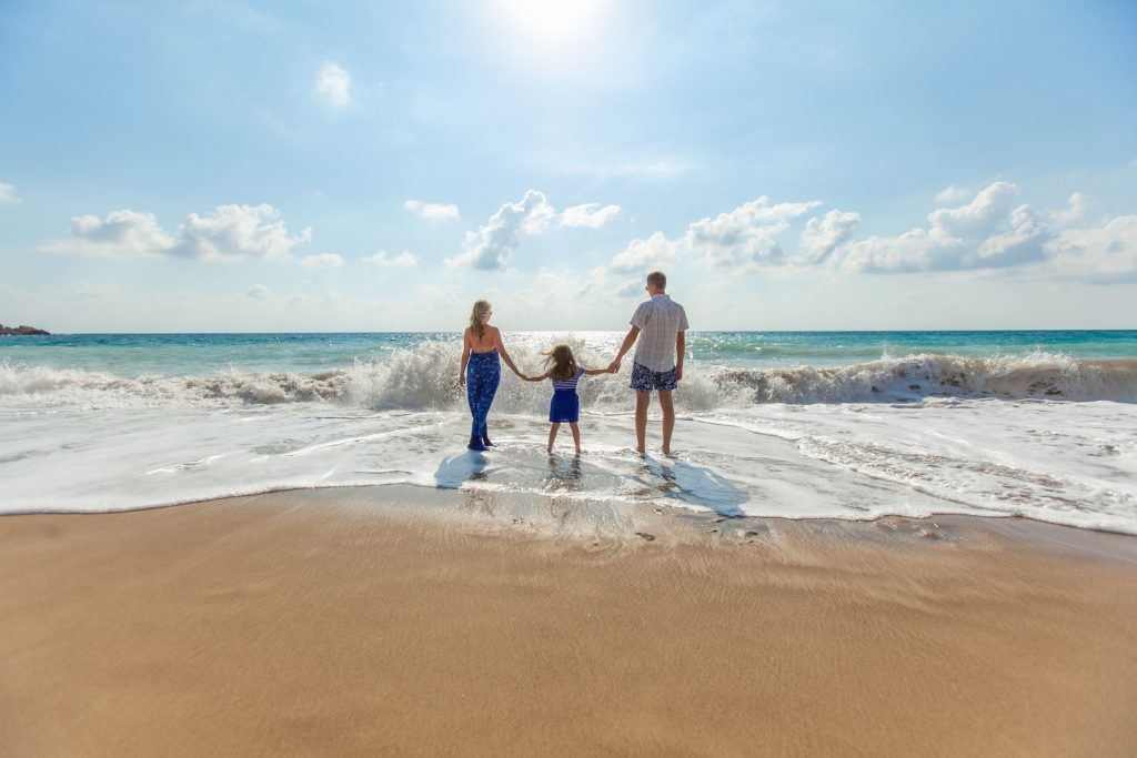 Family enjoying a sunny day at the beach with ocean waves in the background, holding hands and facing the sea